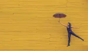 Health help, woman floating with brown umbrella in front of a yellow wooden wall.