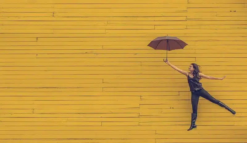 Health help, woman floating with brown umbrella in front of a yellow wooden wall.