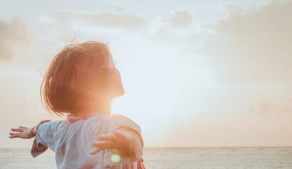 Woman standing in front of a lake with arms open having positive energy.