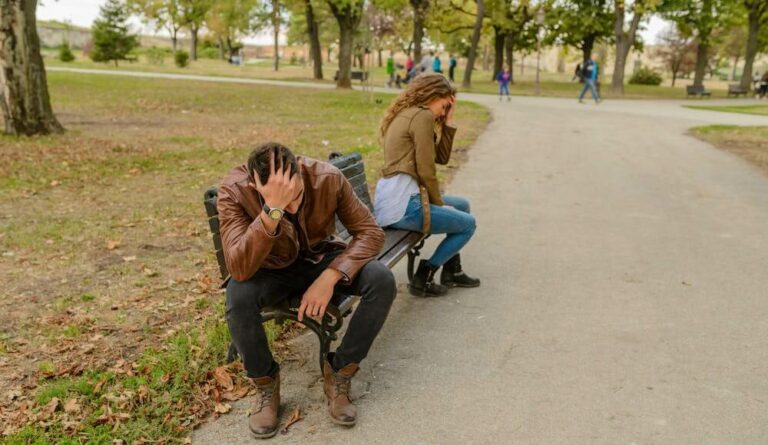 A couple arguing about how to break a love spell, sitting on a bench in a park with their backs to each other.